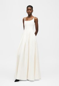 Donna che indossa un abito bianco senza maniche, lungo fino a terra con pieghe, in piedi con le mani dietro la schiena contro uno sfondo chiaro e uniforme.