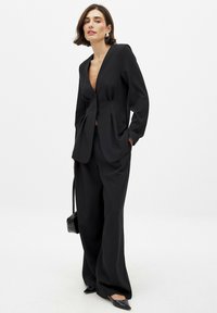 Schwarzer, maßgeschneiderter Anzug mit einem tiefen V-Ausschnitt am Blazer und einem Knoten-Detail, weitem Hosenbein und spitzen Ballerinas. Glatter Stoff und minimalistisches Design.