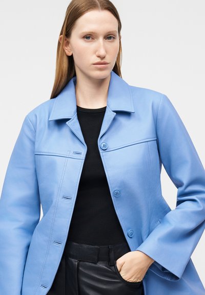 Γυναίκα με καστανά ίσια μαλλιά φοράει ανοιχτό μπλε παλτό με κουμπιά πάνω από μαύρο τοπ και μαύρο δερμάτινο παντελόνι, με το χέρι στην τσέπη.