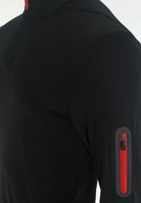 Czarna sportowa bluza z suwakiem do ćwiartki, wyposażona w czerwony zamek błyskawiczny, teksturowaną panel na ramieniu oraz kieszeń na ramieniu z czerwonym zamkiem.