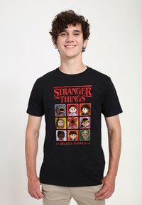 Camiseta de algodón negra con un gráfico de arte píxel de "Stranger Things", que incluye varios personajes en una cuadrícula con el texto "SELECCIONAR JUGADOR".
