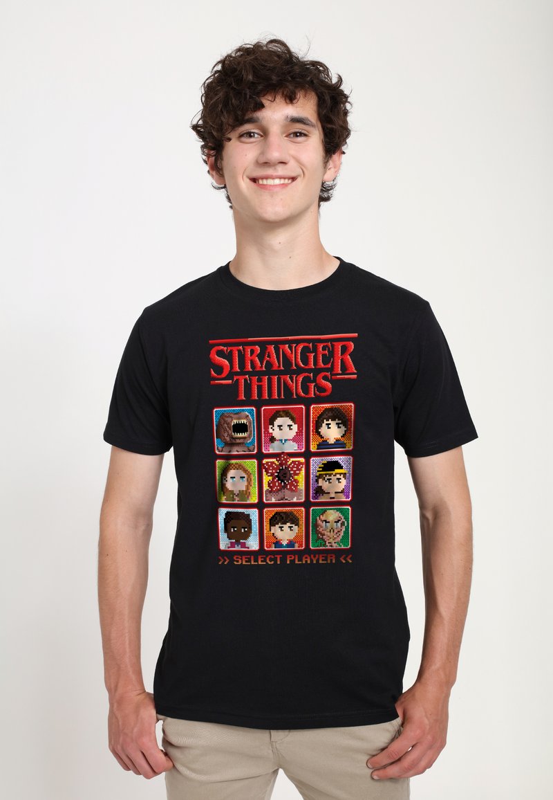 Camiseta de algodón negra con un gráfico de arte píxel de "Stranger Things", que incluye varios personajes en una cuadrícula con el texto "SELECCIONAR JUGADOR".