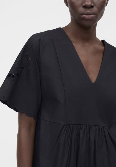 Femme portant une robe noire avec un décolleté en V et des manches courtes festonnées présentant des découpes brodées florales.