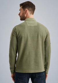 Langarm-Olivgrünes Poloshirt mit gerippter Textur, seitlichen Schlitzen und einem Aufnäher am linken Ärmel. Lockerer Schnitt mit Kragen.