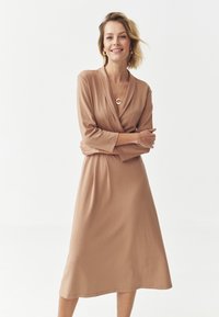 Pitkähihainen, kietaisumallinen beige kangasmekko, jossa on keskipitkä helma ja rento istuvuus. Asusteina on pyöreä kaulakoru.