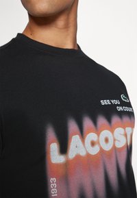 Črna bombažna majica z gradientnim oranžnim in sivim tiskom "LACOSTE", z besedilom "VIDIMO SE NA IGRIŠČU" zgoraj in majhnim logotipom.