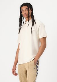 Noormees pikkade punutud juustega, seljas kreemjas lühikeste varrukatega polosärk ja beežid värviliste küljeribadega püksid ning väike logo.