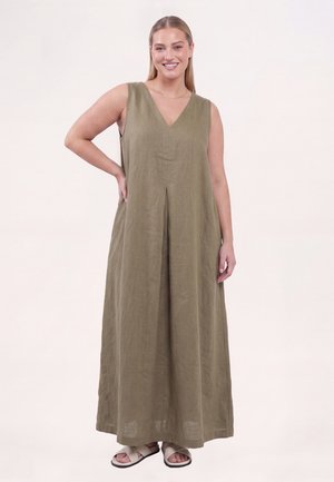 Smilende kvinde med lyst hår iført ærmeløs olivengrøn maxikjole og beige sandaler, stående med den ene hånd på hoften mod ensfarvet baggrund.