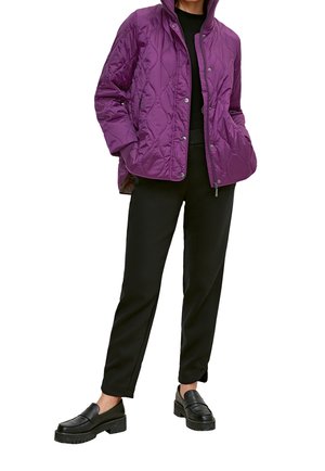 Person iført en lilla quiltet jakke, sort top, sorte bukser og sorte loafers, stående med hænderne i jakkelommerne.