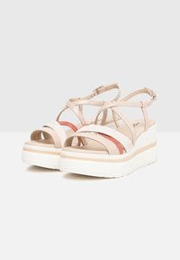 Beige platform sandals with crisscross straps, featuring a coral accent and a white textured sole. Adjustable ankle strap for fit.