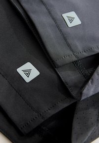 Schwarzer Stoff mit glatter Textur, versehen mit dreieckigen Logo-Patches und genähten Rändern, die ein mehrschichtiges Design mit perforierten Details zeigen.