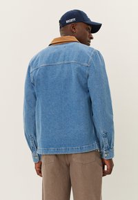 Jeansjacke in Hellblau mit einem braunen Kragen, verfügt über Knopfverschlüsse und einen geraden Schnitt. Kombiniert mit braunen Hosen und einer marineblauen Mütze.