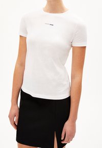 Žena v bílém přiléhavém tričku s nápisem "Trvalá změna" a černé sukni s předním rozparkem.