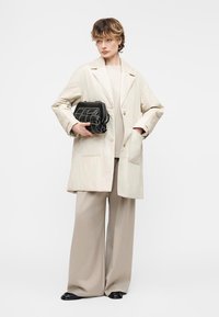 Νεαρό άτομο που φοράει μπεζ υπερμεγέθη παλτό, παντελόνι με φαρδύ πόδι, μαύρα παπούτσια, κρατώντας μια μαύρη υφασμάτινη τσάντα φάκελο σε απλό φόντο.