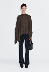 Brauner gestrickter Poncho, lockere Passform, mit weit geschnittenen Ärmeln. Kombiniert mit schwarzen, hochgeschnittenen Hosen und einer kleinen schwarzen Handtasche.