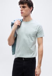 Νέος άνδρας που φοράει ανοιχτό πράσινο κοντομάνικο μπλουζάκι με μικρό λογότυπο στο στήθος, κρατώντας ένα τζιν σακάκι στον ώμο του.