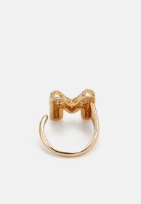 Inel din metal auriu cu un design ondulat, având o formă decupată în forma literei "M" sus. Brățara este netedă și lustruită.