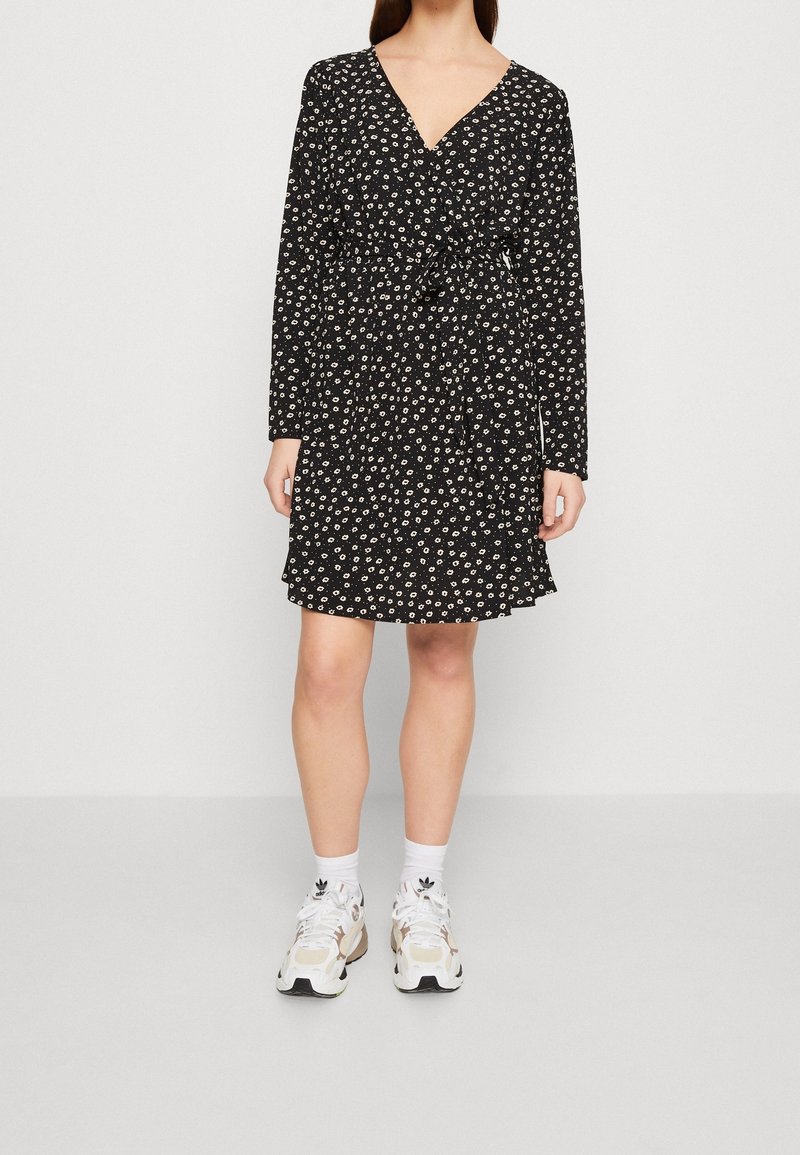 Femme portant une robe noire à manches longues avec imprimé floral blanc, chaussettes blanches et baskets blanches, debout devant un fond clair uni.