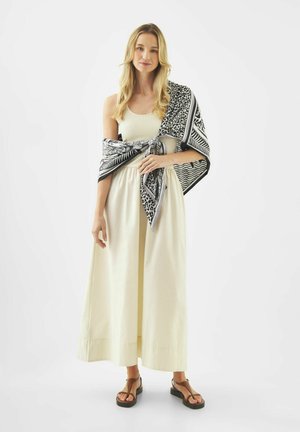 Femme blonde portant une robe maxi crème sans manches et une écharpe à motifs noirs et blancs nouée autour des épaules, avec des sandales noires.