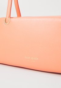 Růžová kožená kabelka s hladkým povrchem, zlatým logem "kate spade NEW YORK" a odpovídajícími rukojetmi, která představuje minimalistický design.