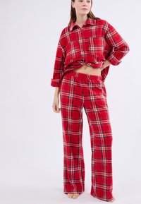 Set di pigiama in flanella a scacchi rossi, composto da una camicia con bottoni e maniche arrotolate e pantaloni con cintura elasticizzata, entrambi con accenti bianchi e neri.