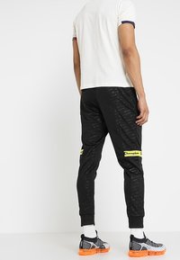 Svarta joggerbyxor med gult "Champion"-logotyp, slim fit, textil med subtila mönster, matchas med vita strumpor och sportiga skor.