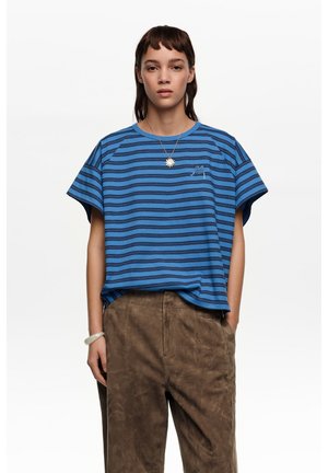 Joven con cabello oscuro de longitud media que lleva una camiseta de rayas azules y negras, pantalones marrones, un collar con forma de sol y una pulsera blanca.