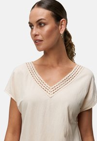 Top color crema con maniche corte e scollo a V, caratterizzato da un motivo intrecciato sul colletto. Tessuto morbido e leggero con una vestibilità comoda.
