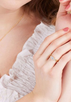 Gouden ring met een centrale ronde steen, omringd door kleinere stenen. De hand van het model rust op rimpelig wit stof, waardoor de schittering van de ring goed zichtbaar is.