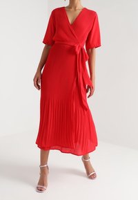 Vestido rojo plisado cruzado con mangas cortas, escote en V profundo, cintura atada y una falda fluida de longitud media. Combinado con tacones de color claro.