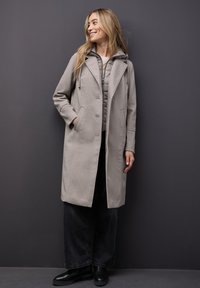 Grau melierter Wollmantel mit Kapuze, seitlichen Taschen und taillierter Passform, über einer gesteppte grauen Innenjacke und schwarzen Hosen getragen.