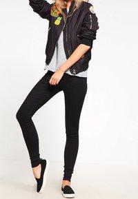 Schwarze Bomberjacke mit Aufnähern, graues T-Shirt darunter, schwarze Leggings und schwarze Slipper. Das Material wirkt leicht und figurbetont.