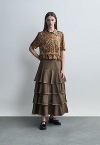 Žena u smeđoj bluzi kratkih rukava s paisley uzorkom i smeđoj višeslojnoj dugoj suknji s ukrasima, stoji uz jednobojni sivi zid.