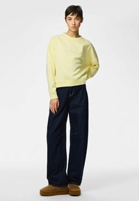 Sudadera amarilla de corte relajado con puños de canalé, combinada con vaqueros de pierna ancha azul oscuro y zapatillas marrones.