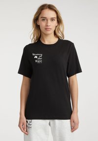 Sort bomulds T-shirt med korte ærmer og rund hals. Har hvid tekst, der siger "Women of the Wave" på venstre bryst.