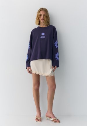 Mlada žena nosi mornarsko plavu majicu dugih rukava s plavim printovima sunca i tekstom, krem mini suknju s čipkama i bež sandale s potpeticama.