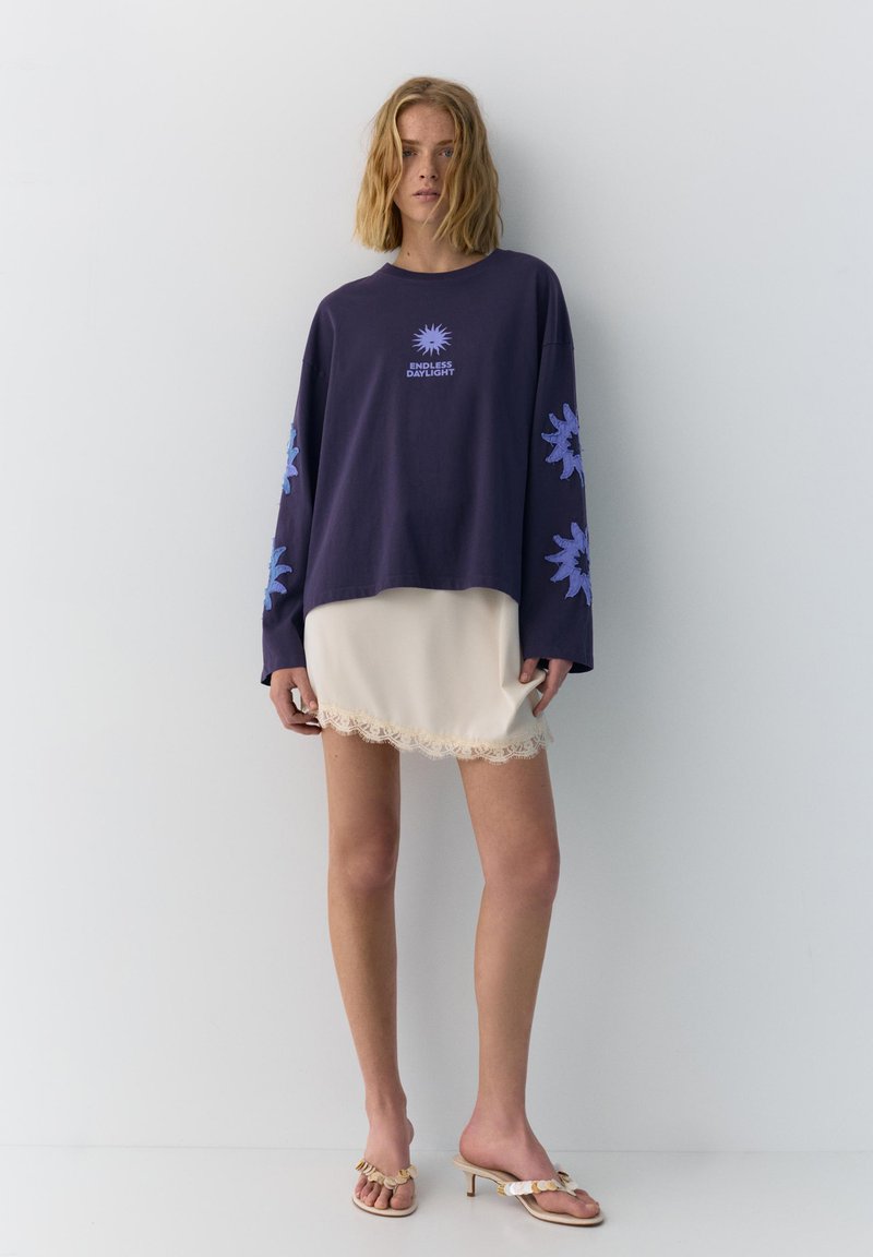 Jeune femme portant un haut à manches longues bleu marine avec des imprimés de soleil bleus et du texte, une mini-jupe crème avec bordure en dentelle et des sandales beiges à talons.