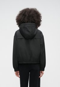 Osoba s kučeravými vlasmi, oblečená v čiernej kapucňovej bunde a čiernych nohaviciach, stojaca chrbtom k nám pred jednoduchým svetlým pozadím.