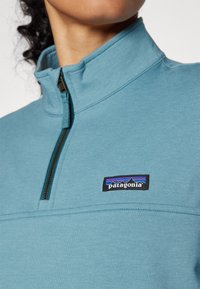 Hellblauer Fleece-Pullover mit hohem Kragen und halbem Reißverschluss. Mit einem schwarzen Patagonia-Logo-Patch auf der Brust. Glatte Textur.