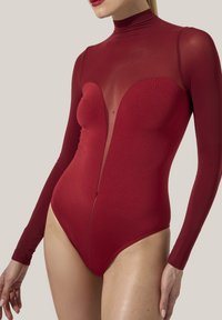 Kvinna iklädd en åtsittande långärmad, genomskinlig röd bodysuit med solida och transparenta tygpaneler på bålen och armarna.