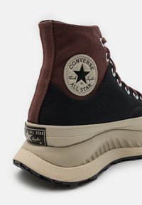 Vysoké tenisky s hnědým plátnovým svrškem a černou spodní částí, vybavené kulatou nášivkou s logem a silnou béžovou gumovou podrážkou.