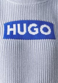 Gebreide trui in lichtgrijze wol met een ribbeltextuur. Bevat een gedurfde blauwe horizontale band met "HUGO" in witte letters.