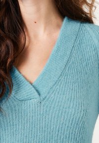 Helles blaues, strukturiertes Strickpullover mit tiefem V-Ausschnitt und ribbeligen Kanten. Das Material wirkt weich und gemütlich und zeigt feine Fadendetails.