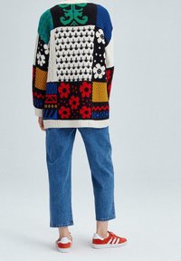 Pull multicolore en tricot avec des motifs géométriques et floraux, associé à un jean bleu clair et des baskets rouges avec des accents blancs.