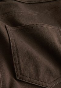 Detailní záběr hnědé látky zobrazující čtvercovou kapsu s viditelným šitím a texturovaným vzorem tkaní.