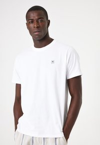 Camiseta de algodón blanca con cuello redondo, mangas cortas y un pequeño logo cuadrado en el lado izquierdo del pecho. Textura suave, ajuste estándar.