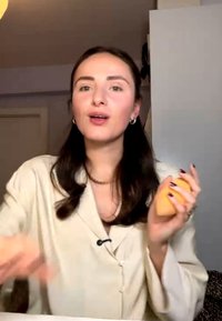 Jeune femme aux longs cheveux foncés tenant une éponge à maquillage orange, portant une veste claire, parlant à l'intérieur à un bureau.