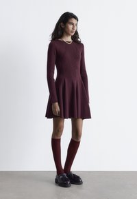 Burgunderfarbendes, geripptes Strickkleid mit langen Ärmeln und weit ausgestelltem Rock, kombiniert mit knielangen burgunderfarbenen Socken und schwarzen Tassenschuhen.