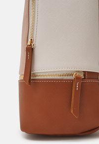 Gros plan d'une section beige et marron texturée de sac avec deux fermetures éclair dorées visibles et des tirettes en cuir marron.