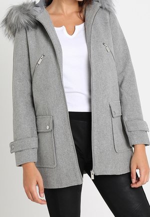 Femme portant un manteau d'hiver gris avec une capuche bordée de fourrure, une fermeture éclair devant, des poches à rabat et des poignets boutonnés, par-dessus un haut blanc et un pantalon noir.